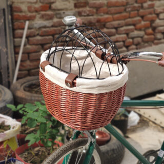On voit la photo d'un panier pour chien à l'avant d'un vélo. Il est en rotin et doublé d'un tissu crème. Le haut du panier possède un couvercle en acier, pour éviter que le petit chien ne tombe. Le vélo est vert et présenté devant un mur de brique.