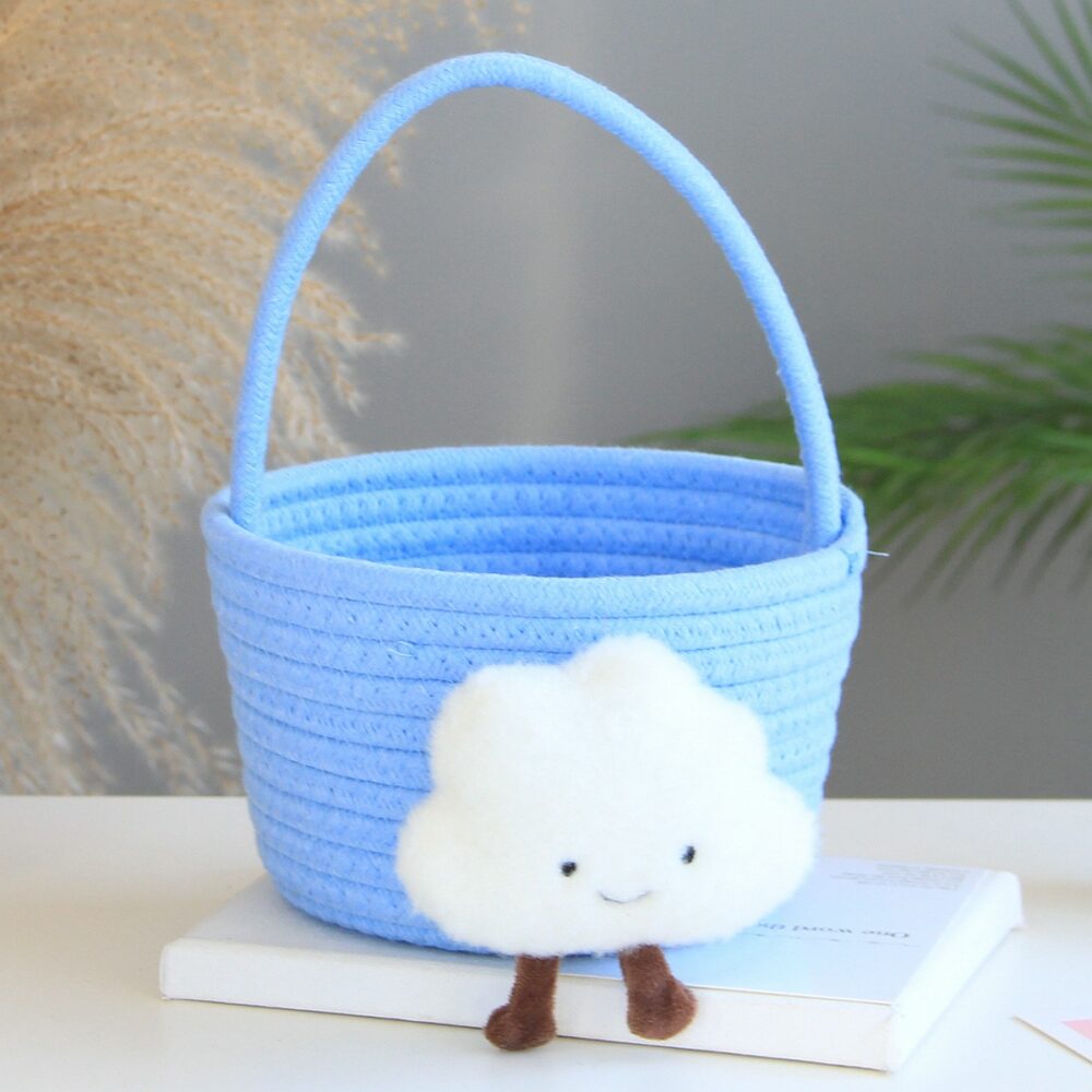 petit panier de rangement pour enfants bleu avec un doudou de nuage blanc, avec des yeux, un sourire et des jambes marrons qui pendent, sur un livre fin blanc, qui est sur une table blanche, avec une plante verte partiellement visible derrière