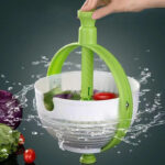 Main poussant un manche vert qui fait tourner un panier à salade blanc avec des fruits dedans avec des gouttes d'eau qui sortent du panier