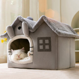Dans une pièce avec de la moquette, on voit un panier pour chat en peluche en forme de maisonnette. il est gris et blanc. A l'intérieur, il y a un chat blanc persan qui dort.