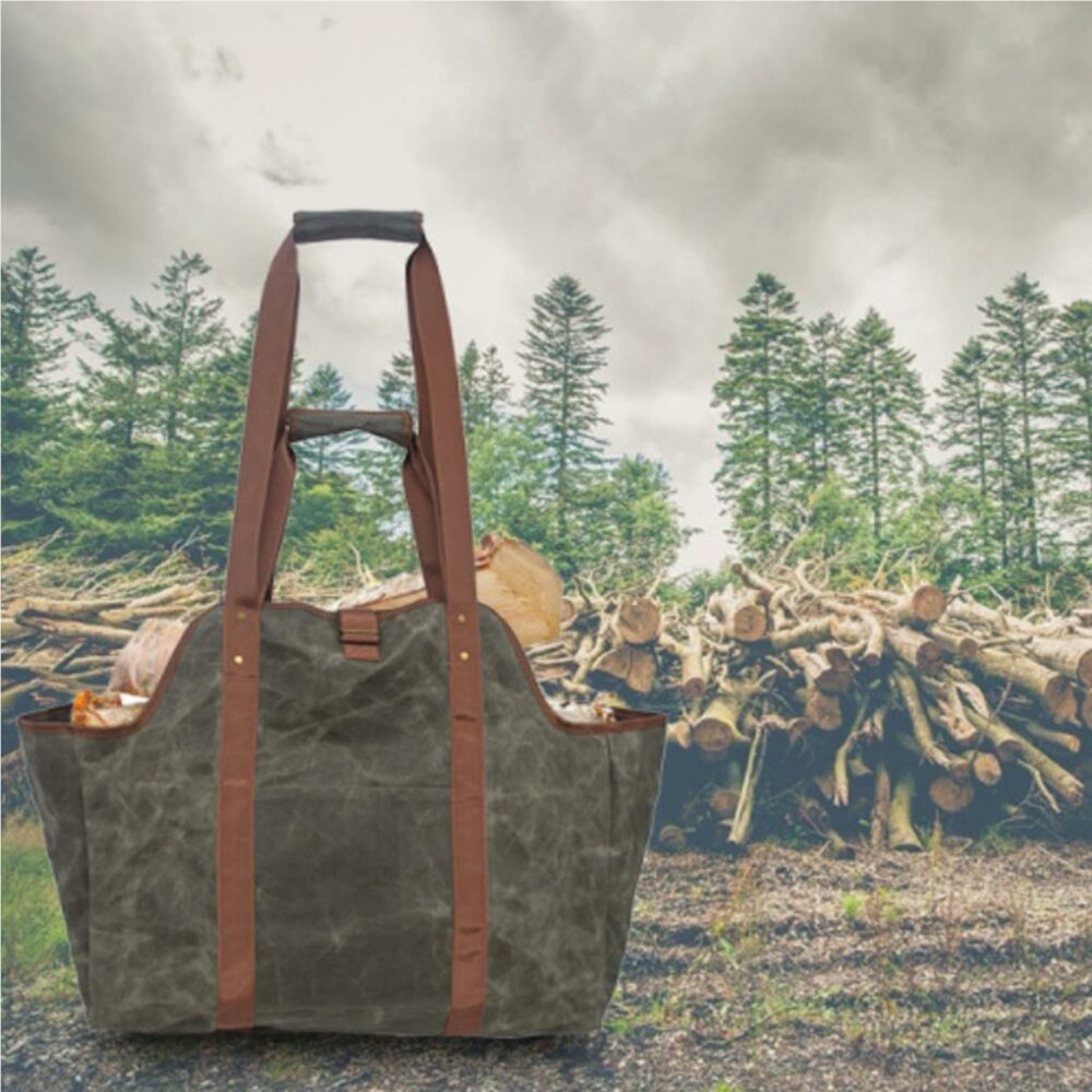 En premier plan , un grand sac gris-vert avec grandes anses marrons avec des buches de bois à l'intérieur. Il y a un tas de bois coupé en arrière plan avec une forêt et un ciel gris