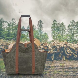 En premier plan , un grand sac gris-vert avec grandes anses marrons avec des buches de bois à l'intérieur. Il y a un tas de bois coupé en arrière plan avec une forêt et un ciel gris
