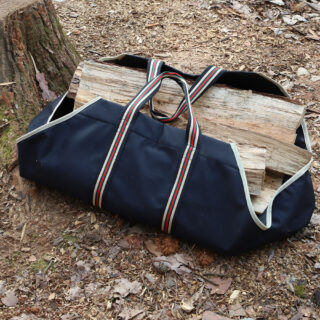 Sur un sol de forêt près d'un tronc d'arbre coupé, un grand sac en tissu bleu avec grandes anses rayées blanches et rouges avec des buches de bois à l'intérieur.