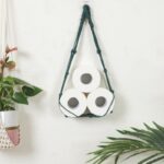 panier mural vert en corde de coton et avec un support en bois, rempli de 3 rouleaux de panier-toilette, attaché sur un mur gris clair, avec une plante en pot suspendu coupée à gauche de l'image, et une tige d'une autre plante en bas à droite