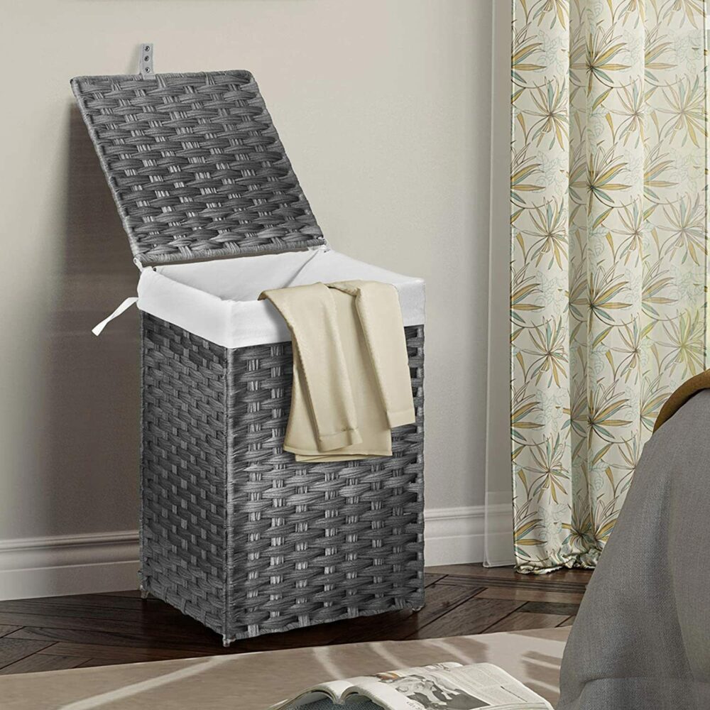 7838-j9oukh.jpg Panier à linge gris avec un couvercle ouvert et du linge qui dépasse. Le panier se trouve dans une pièce avec un rideau derrière
