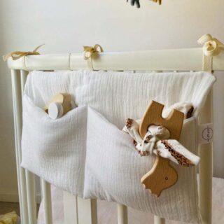 Panier à deux compartiments en coton blanc accroché par des lanières au lit à barreaux en bois d'un bébé avec des petits jouets en bois à l'intérieur